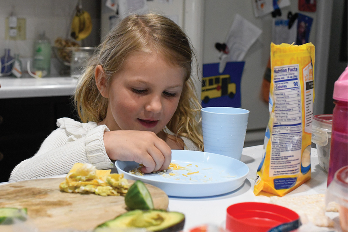 Niña en una mesa de cocina comiendo una comida con aguacate, papas fritas y jugo en la mesa.