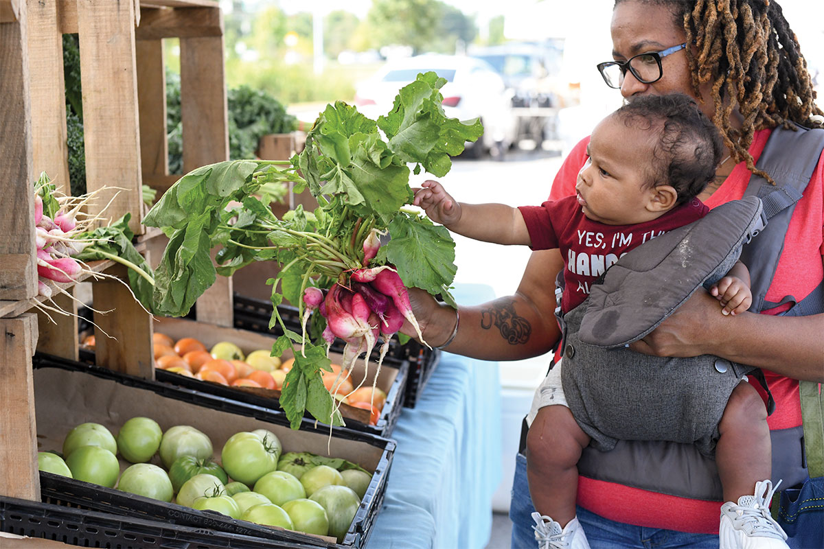 Mujer sosteniendo a un bebé en una mochila portabebés en su parte delantera mientras recoge rábanos frescos en un mercado agrícola al aire libre.
